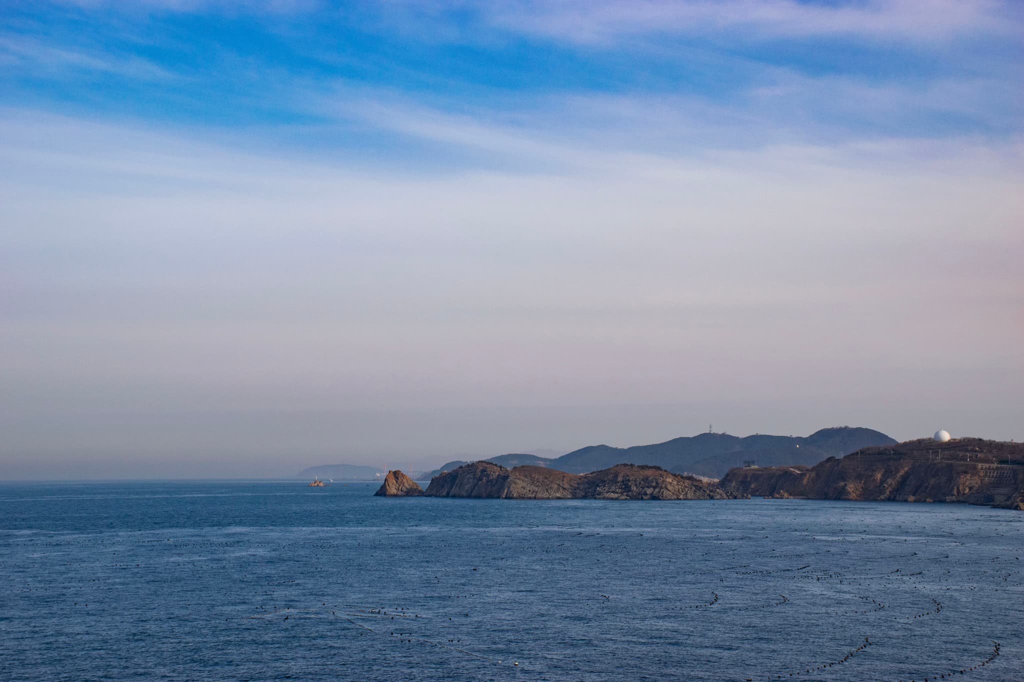 海面平静,远处的山峦在天边若隐若现,天空晴朗,微带云彩。